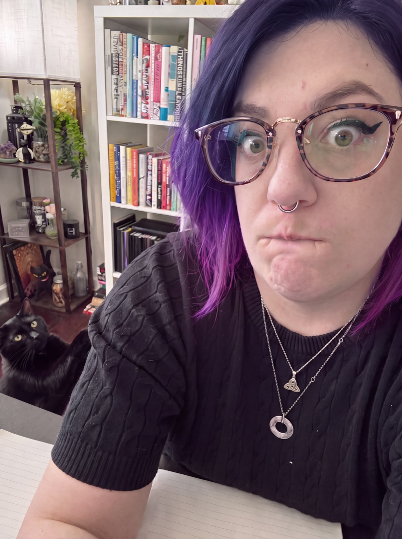 Theresa at her desk, making a skeptical face at the camera, with her black cat Carmy behind her.